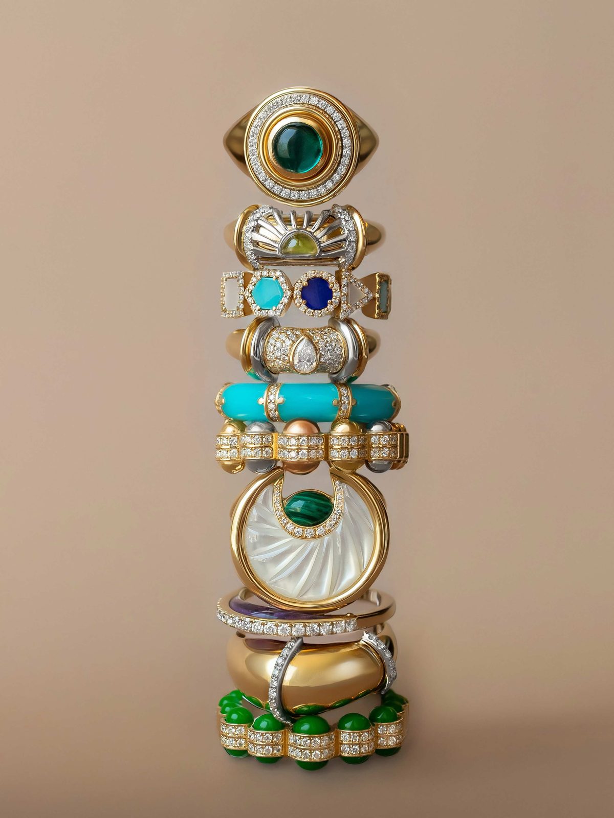 A stack of rings balanced one on another — a green cabochon eye signet, a turquoise bar, a pavé band, a carved mother-of-pearl disc and a row of jade beads.