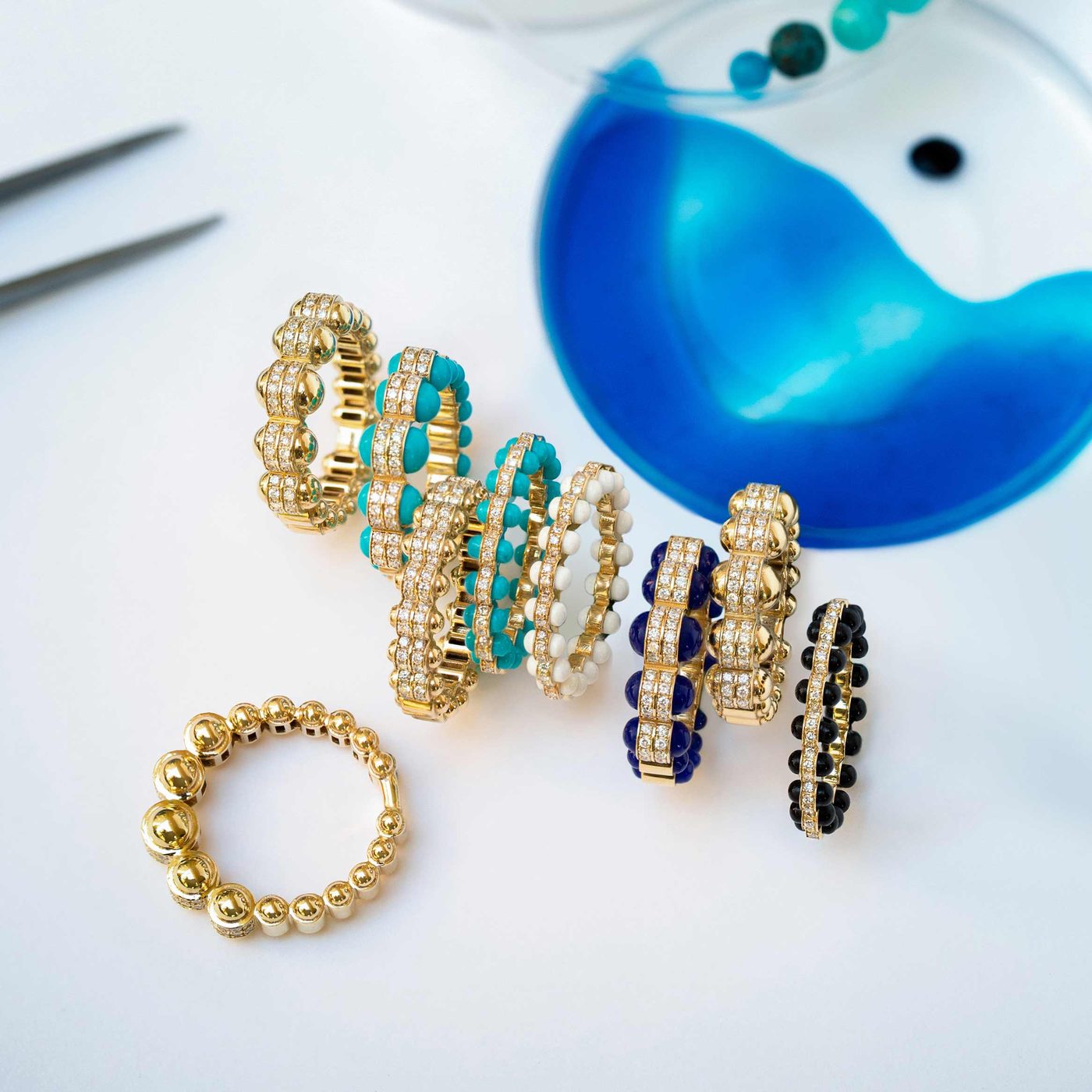 Atoms rings tumbled on a white bench beside a blue agate dish — beaded gold bands set with diamonds and rows of turquoise, white, lapis and onyx beads.