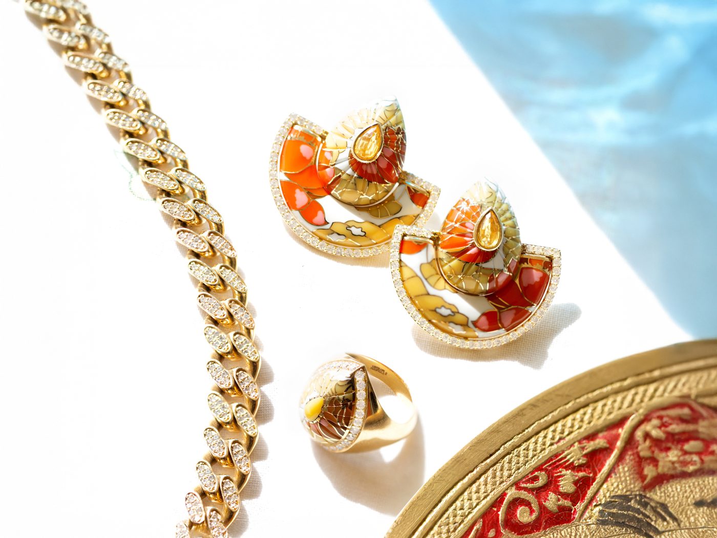 A diamond-set gold curb chain beside enamelled fan earrings and a matching dome ring, photographed in daylight against gilded metalwork.