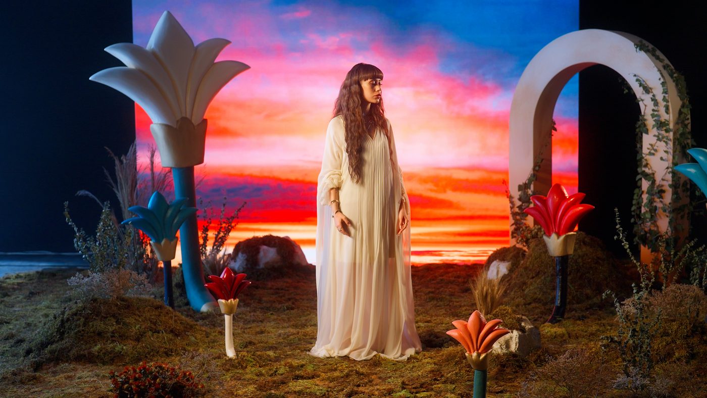 A Psychedeliah campaign still: a model in a cream kaftan standing in a painted sunset among oversized sculpted flowers.