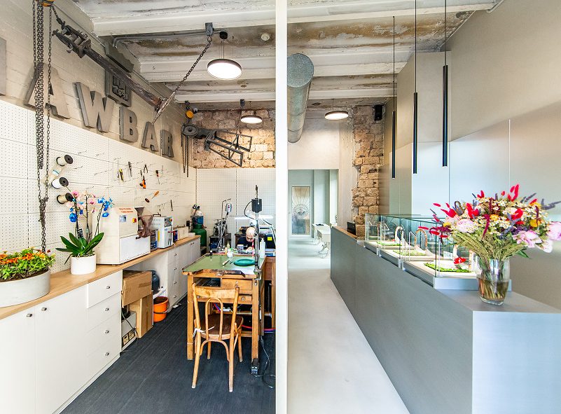 Inside the Beirut atelier: a jeweller at the workbench under the NAWBAR letters, beside the boutique's lit vitrines and a vase of wildflowers.